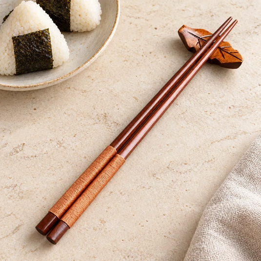 Baguettes japonaises en bois foncé à cordon beige sur un repose-baguettes en forme de feuille, près d’une assiette d’onigiri.