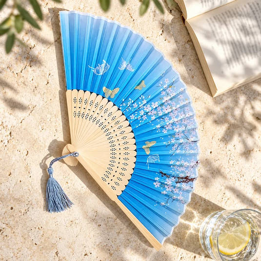 Éventail japonais Chōchō bleu posé sur une table de terrasse, près d’un livre et d’un verre d’eau citronnée.