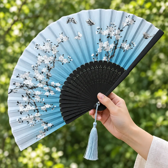 Éventail japonais Ume bleu tenu en main dans un jardin, avec fleurs blanches et pompon bleu clair.