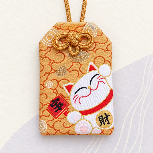 Omamori japonais doré orangé, brodé d’un maneki-neko blanc et de motifs de pièces.