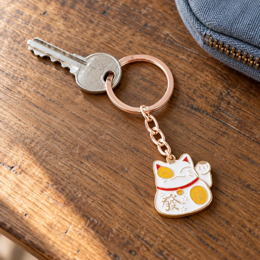 Porte-clés Maneki Neko blanc relié à une clé sur son anneau doré rosé, posé sur une table en bois.