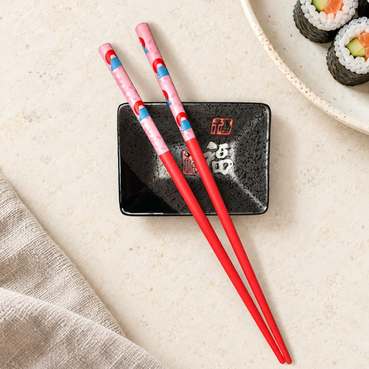 Baguettes japonaises Fuji-San Aka rouges sur une coupelle noire, près d’une assiette de maki.
