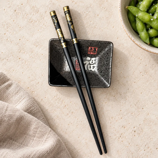 Baguettes japonaises Oni Kuro noires et dorées sur une coupelle noire, près d’un bol d’edamame.