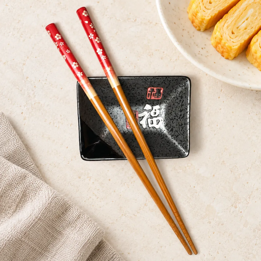 Baguettes japonaises Sakura Aka à manches rouges sur une coupelle noire, près d’une assiette d’omelette japonaise.