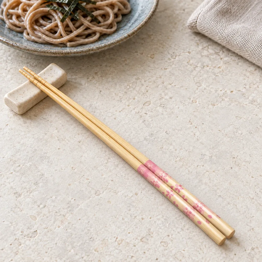 Baguettes japonaises Sakurairo à fleurs roses, posées sur un repose-baguettes près d’une assiette de soba.