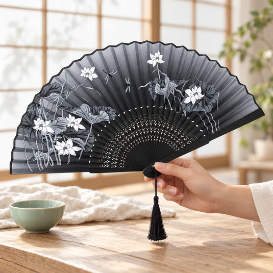 Éventail japonais noir et gris à fleurs et libellules tenu en main près d’un bol en céramique vert clair.