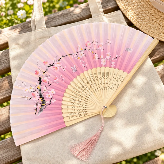 Éventail japonais Sakura rose posé sur un sac en toile, près d’un chapeau de paille sur un banc.