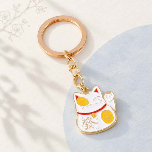 Porte-clés japonais Maneki Neko blanc, avec patte levée, détails jaunes, collier rouge et chaîne dorée.